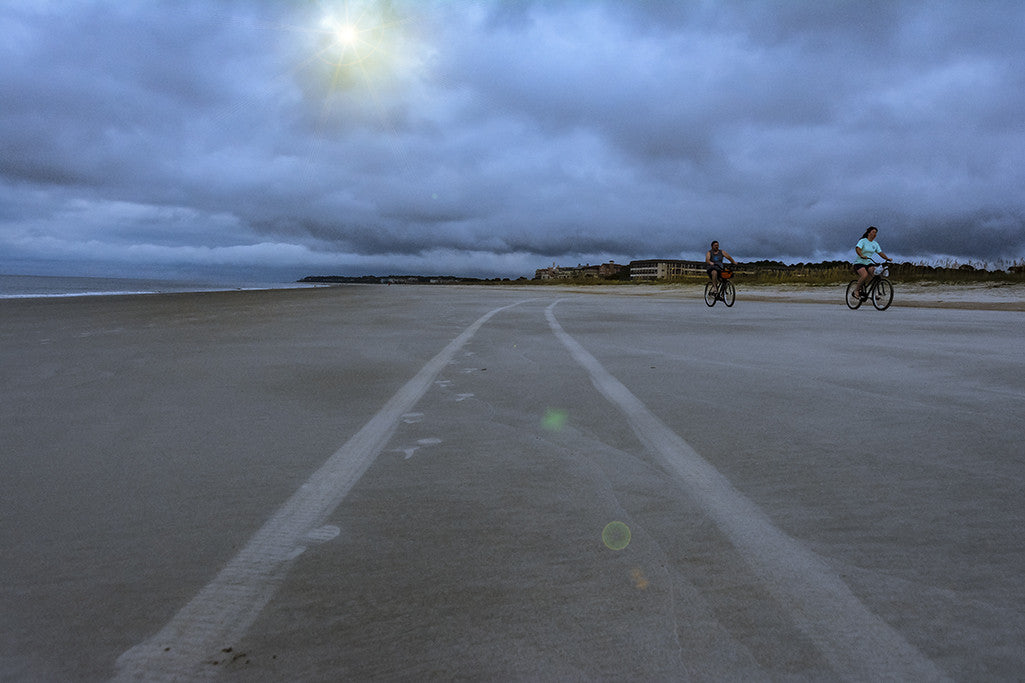 Bikers ride the beach on Hilton Head Island under an overcast but colorful sky.